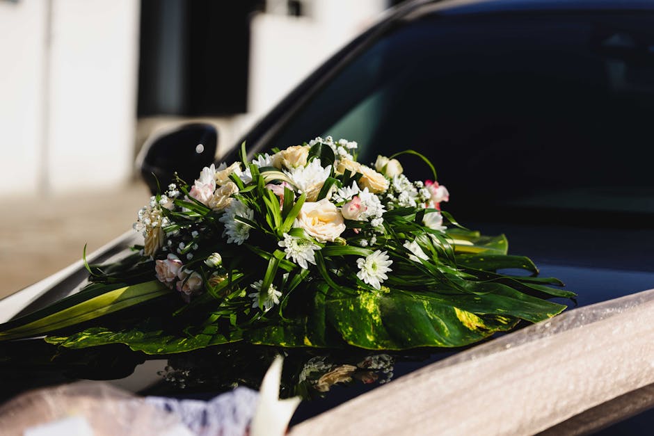 A floral arrangement placed on the hood of a black car, featuring a bouquet of fresh white and blush pink flowers, including roses, daisies, and miniature filler flowers, accented with lush green foli