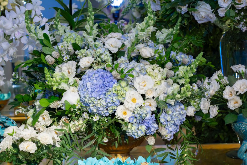 A large floral arrangement featuring clusters of light blue and white hydrangeas, surrounded by white lisianthus, small white daisies, and various green foliage, including broad leaves and sprigs. The
