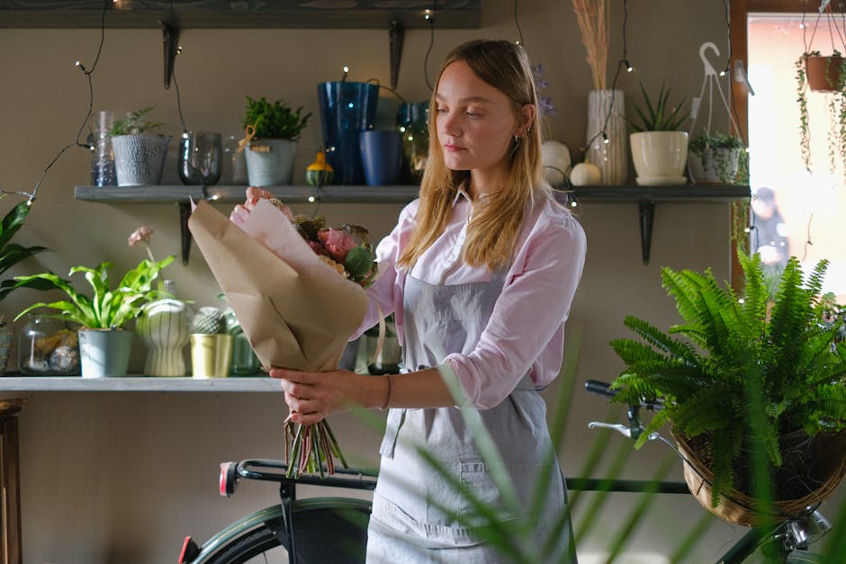 A young woman with long, light brown hair, dressed in a pale pink shirt and light grey apron, is holding a bouquet of fresh flowers wrapped in brown paper. The bouquet includes pink roses, white bloss