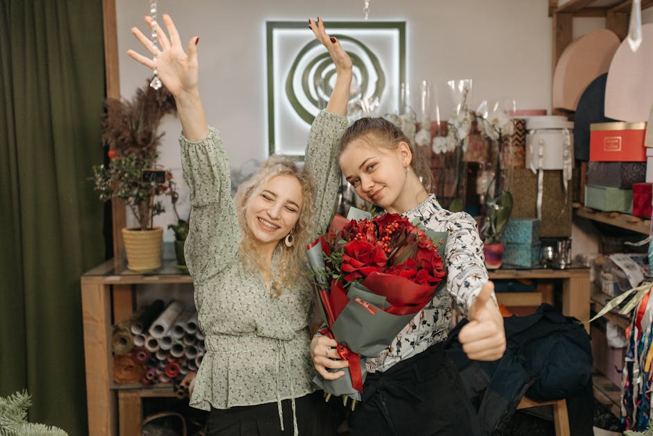 Two young women in a florist's workshop, one with curly blonde hair smiling broadly with arms raised, and the other with straight brown hair holding a large bouquet of vibrant red roses wrapped in gra