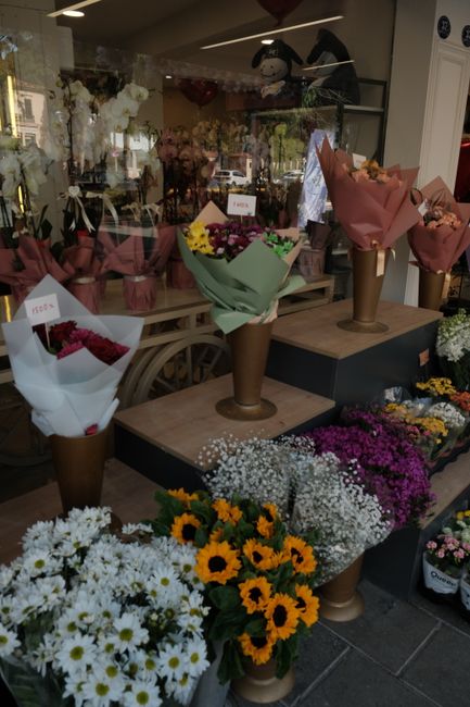 A display of fresh flower bouquets arranged in tall, cylindrical brown paper wrappers with pink and green wrapping paper accents, set on a wooden and black display stand inside a flower shop. The bouq