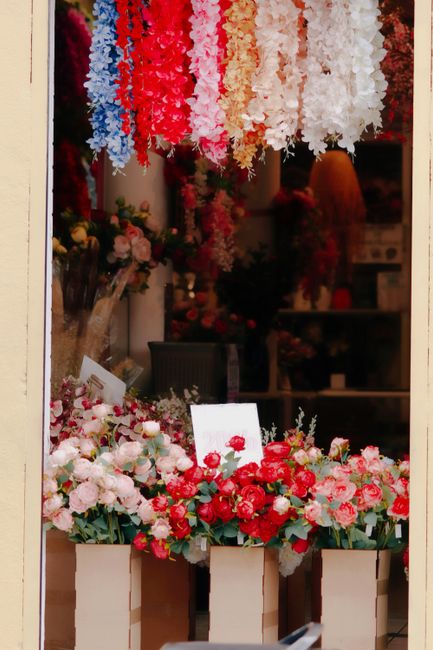A floral display in a flower shop featuring a vibrant arrangement of fresh roses, peonies, and other blooms in shades of pink, red, and white, arranged in white vase-like containers on a wooden surfac