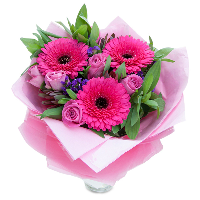 I picture this bouquet sitting on a sunny windowsill near Brentwood Common, its colours both playful and refined. Three vivid fuchsia Gerbera daisies open boldly at the heart, their many petals forming striking, sunflower-like faces around dark centres that provide a dramatic contrast. Around those focal blooms nestle several small roses in a soft lavender-pink or dusty rose tone; their petals are still tightly wrapped in the fresh-bud stage, promising a slow unfurling of tenderness. Little clusters of purple statice or limonium are tucked into the spaces between, lending an airy, almost wildflower quality that breaks the formal lines with gentle whimsy. Generous sprigs of deep green foliage-broad leaves reminiscent of salal or eucalyptus-frame and lift the composition, offering a cool backdrop that accentuates the pinks. The wrapping is deliberately layered: a matte, pale pink tissue paper inside cradles the stems, while a translucent fuchsia-pink cellophane, slightly crinkled, adds a soft shimmer and completes the monochrome story. The arrangement is finished so you can see the top rim of a clear glass vase beneath the folds, ready for immediate placement by a bedside or on a hallway console after a walk from Weald Country Park. In the quiet light the petals hold tiny highlights, the scent leans gently toward rose-scented sweetness with a hint of green from the gerberas, and the overall effect is intimate and celebratory-suitable for a birthday, an anniversary, or the simple kindness of sending flowers to cheer a neighbour in Brentwood or a friend catching the train at Shenfield station.